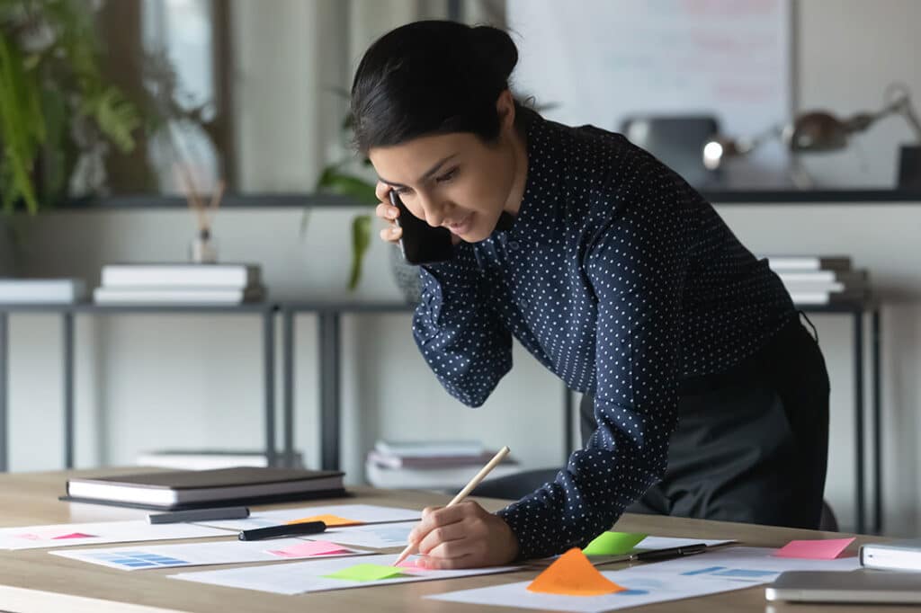 Indian businesswoman talking on cellphone, writing notes on colorful sticky papers in office in career lattice