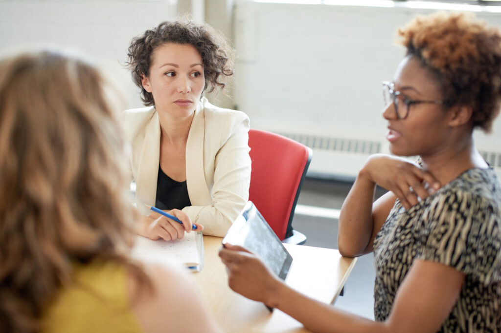 Three business women sitting at a conference table confronting their boss