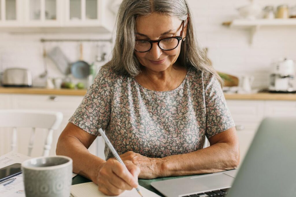 Photograph of a mature white woman with long grey hair and black thick rimmed glasses looking down and doing a personal swat analysis at her kitchen table.