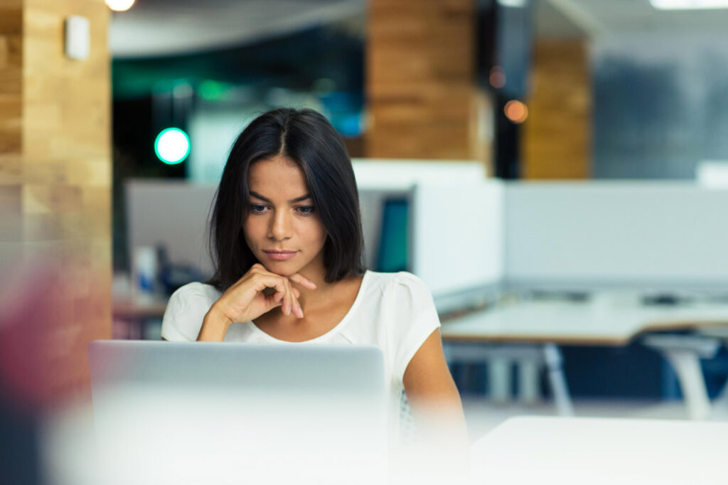 Photograph of a woman with long straight brown hair in a white t-shirt looking intently at a computer screen taking an online professional certificate program