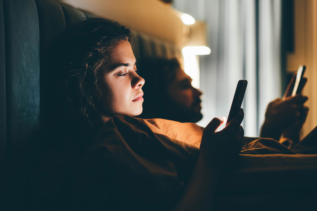 young woman holding mobile phone in bed at night with light shining on face