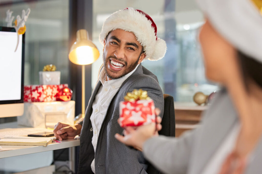 Happy male employee wearing a Santa Claus hat giving his coworker the best ten dollar gift ideas